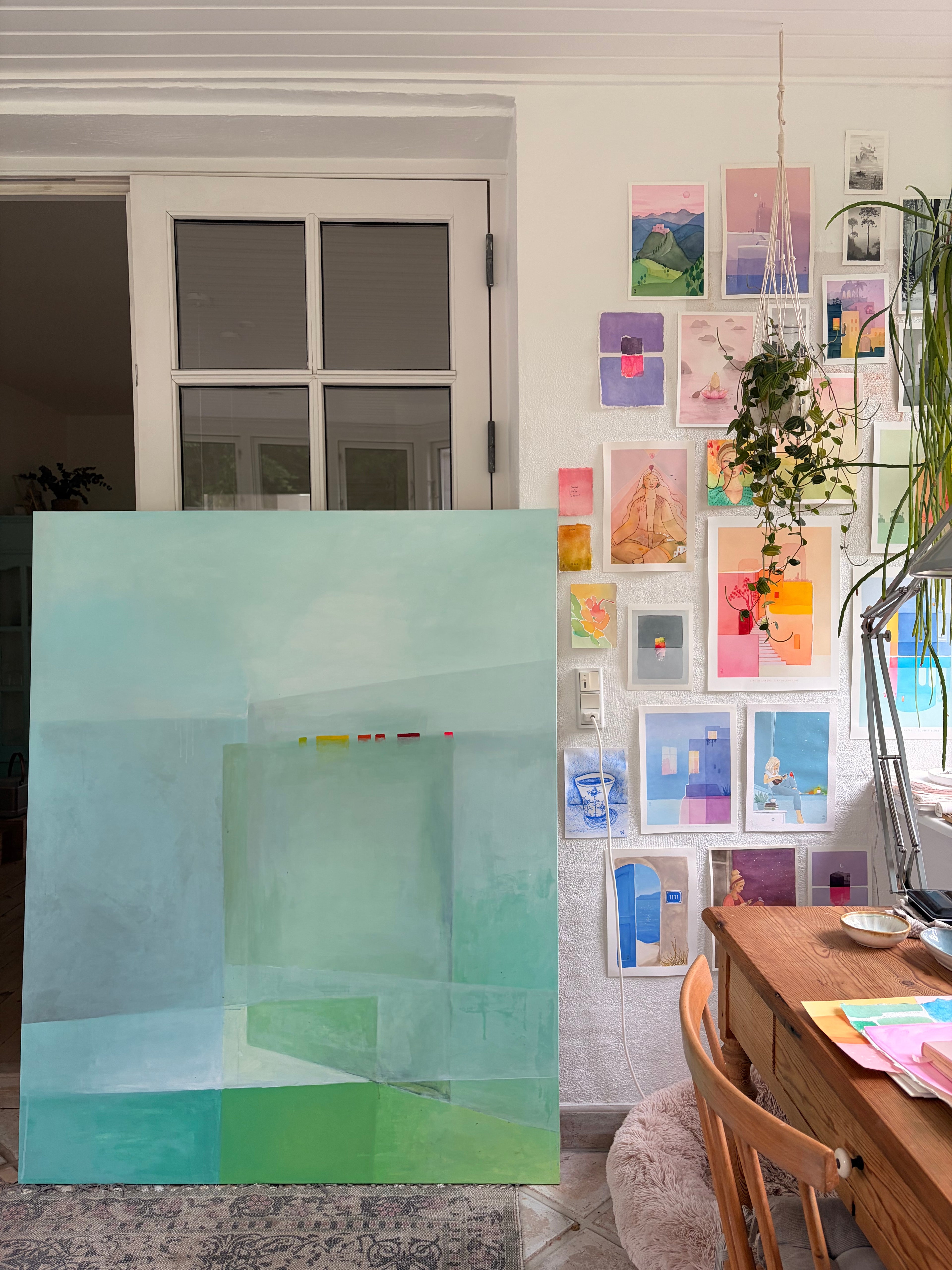 A large abstract canvas painting with soft mint green, aqua, and teal geometric blocks is displayed in a bright room next to a wall covered in colorful framed art prints. A wooden desk and chair are visible in the foreground.