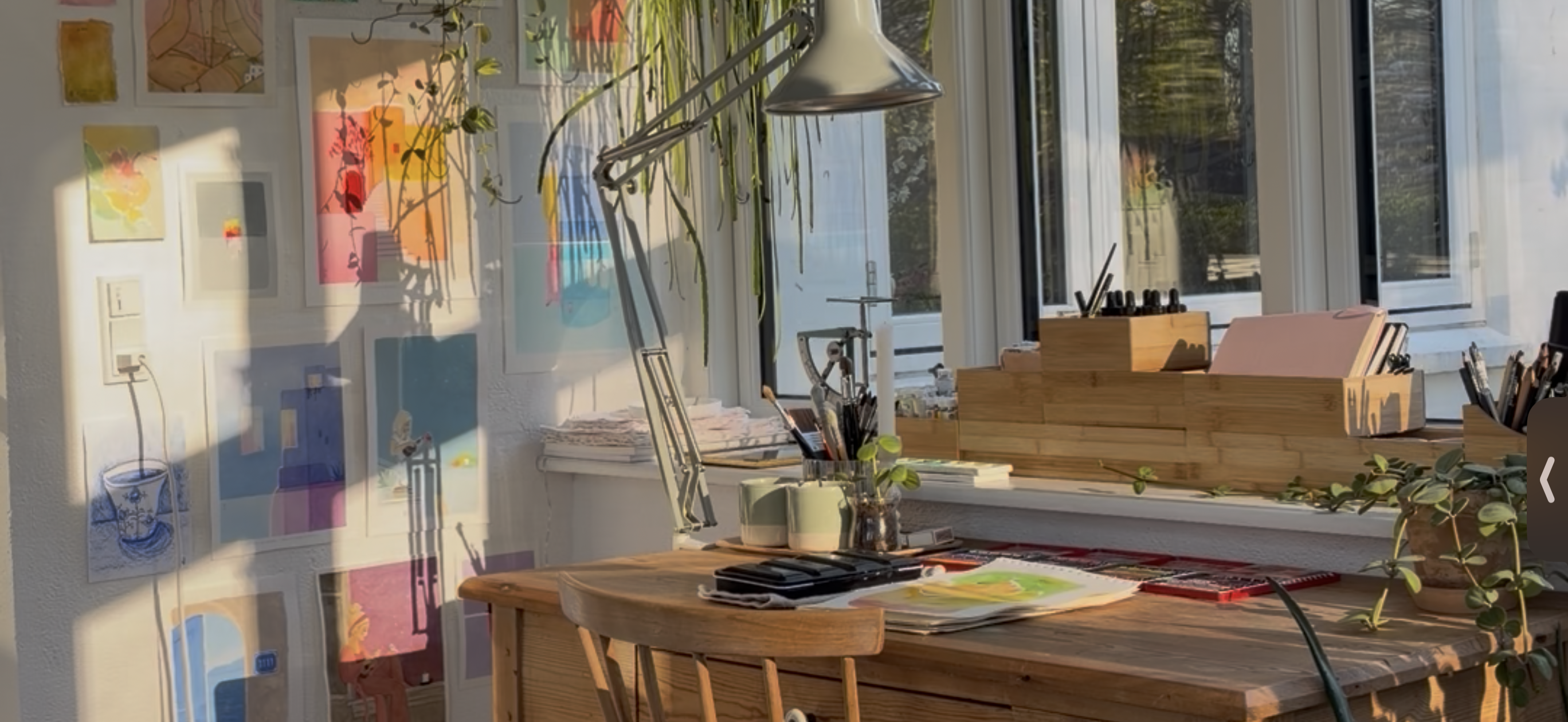 A bright, sunlit art studio or home office with colorful framed artwork on the walls, a wooden desk, a tall floor lamp, and plants near large windows.