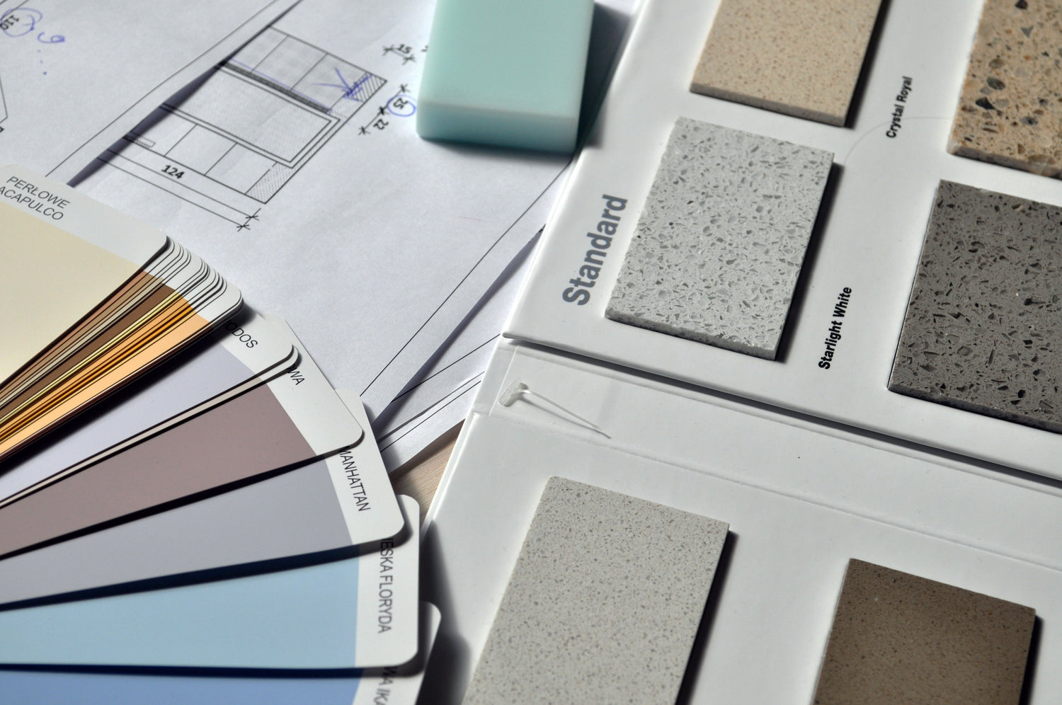 A close-up of a material and color swatch display with multiple rectangular sample tiles in neutral and speckled finishes, alongside printed design sheets and additional color cards.
