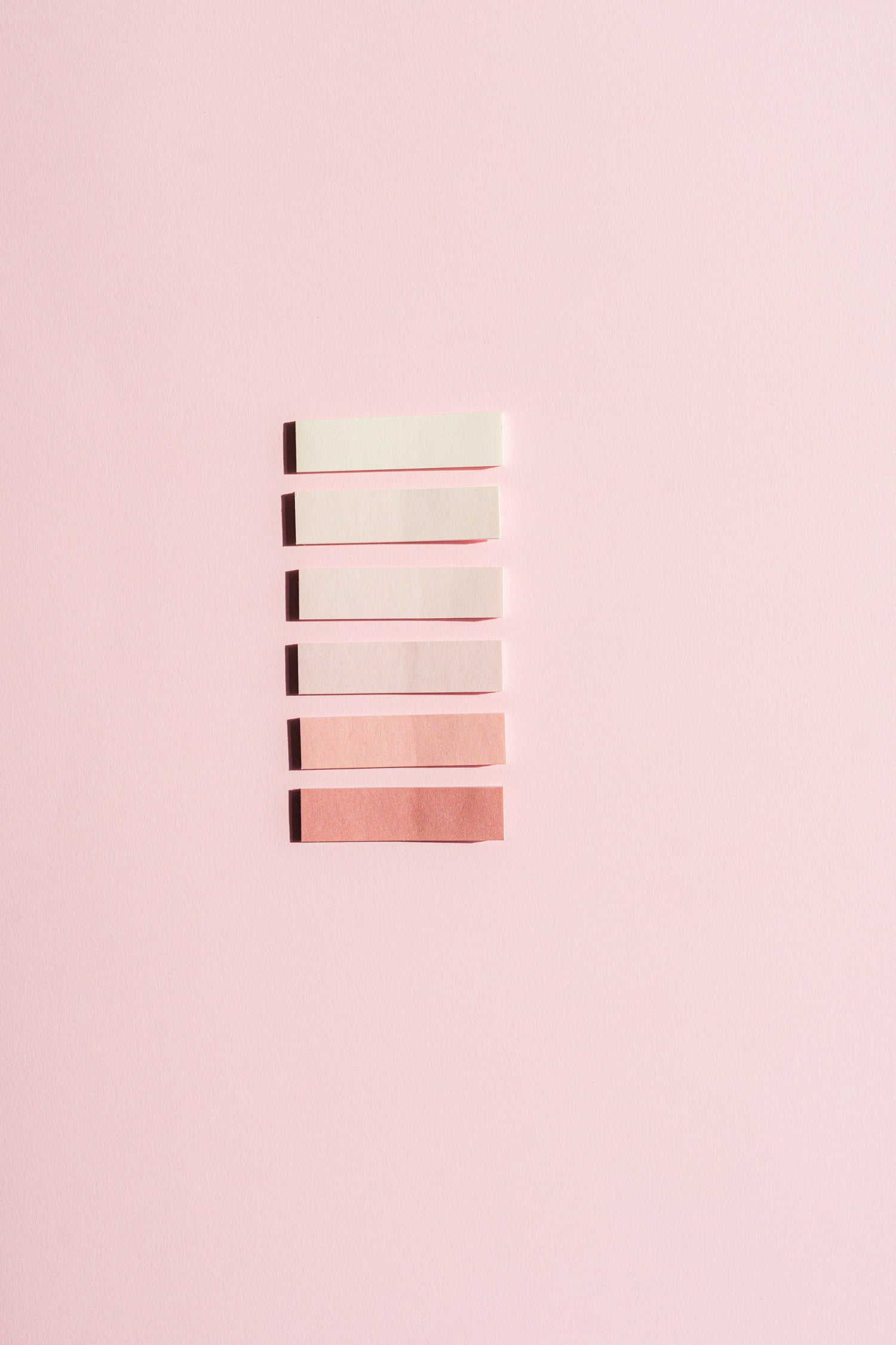 Six rectangular makeup swatches in a vertical stack, showing a gradient from pale cream to light pink and dusty rose, set against a solid light pink background.